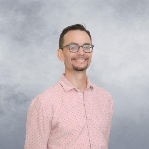 Adam stands for his portrait wearing glasses and a pink shirt.