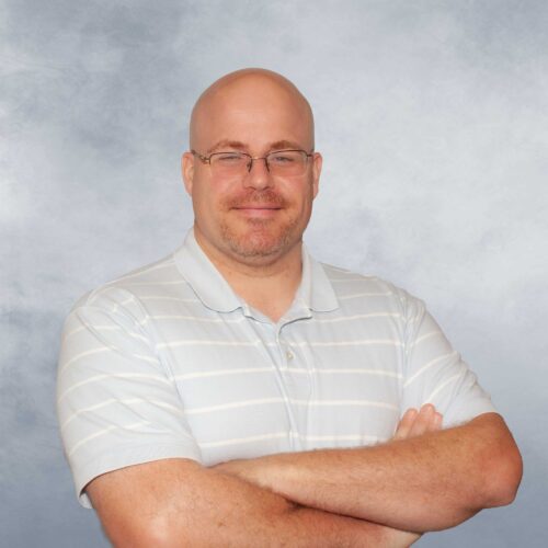 John poses for photo, arms crossed in front of a heathered periwinkle backdrop.