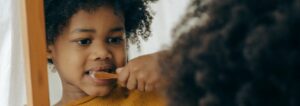 A child in a yellow sweater brushes teeth in front of a mirror