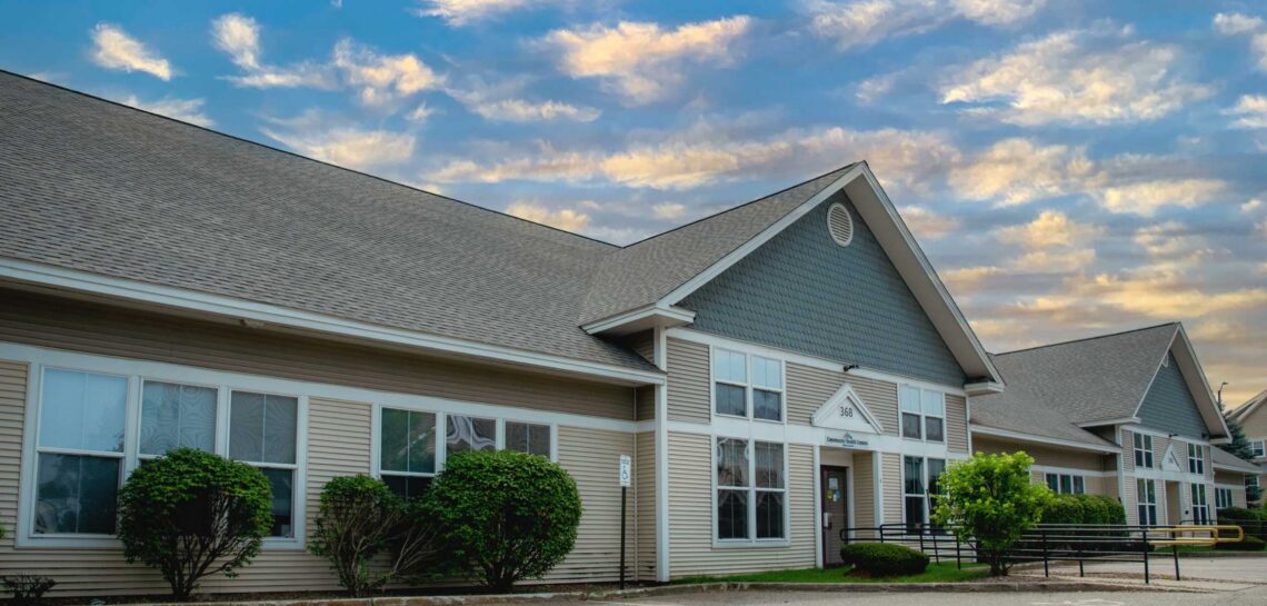 Exterior view of a Good Health Community Health Center location, featuring a large one-story building with beige siding, blue-gray gabled roofs, large windows, landscaped shrubs, and an accessible entrance with ramps under a partly cloudy sky.